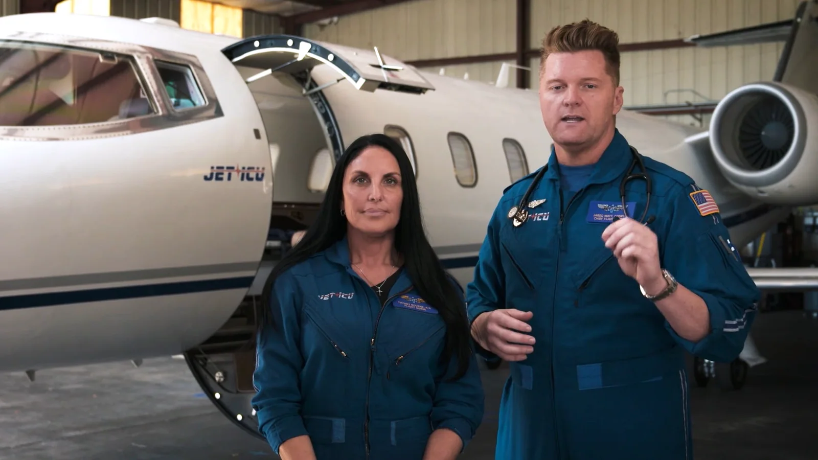Two Jet ICU flight crew in blue flight suits in front of their Learjet in the hangar. (a still from their own video)